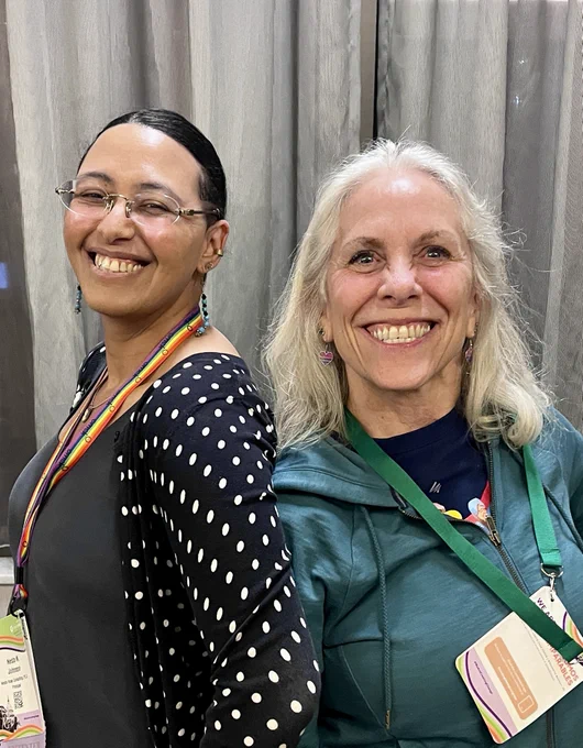 Two people stand back-to-back, smiling warmly at the camera. The person on the left wears glasses, blue beaded earrings, and a black-and-white polka-dot cardigan over a grey top, with a rainbow pride lanyard. Robyn Ochs, on the right, has long, wavy grey hair and wears heart-shaped bisexual pride earrings and a teal zip-up hoodie. Both individuals are wearing event badges clipped to lanyards.