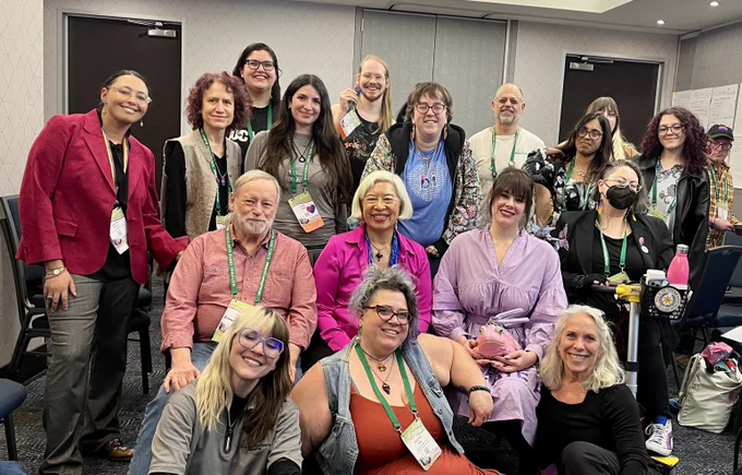 A group photo of approximately 20 people of diverse ages, genders, and backgrounds gathered in a conference room. Most are smiling and wearing colorful lanyards and conference badges. Robyn Ochs is seated in the front row on the far right, wearing a black long-sleeve shirt and smiling. The group is posed closely together, with some individuals standing in the back and others seated in chairs or on the floor in the front.