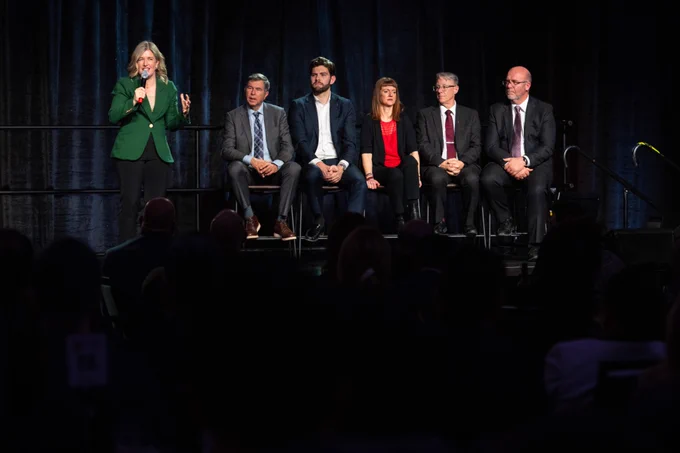 A woman in a green blazer speaks into a microphone on stage while five people, three men and two women in business attire, sit on stools beside her against a dark backdrop.
--
Une femme vêtue d'un blazer vert parle dans un micro sur scène tandis que cinq personnes, trois hommes et deux femmes en tenue de travail, sont assises sur des tabourets à côté d'elle, sur une toile de fond sombre.