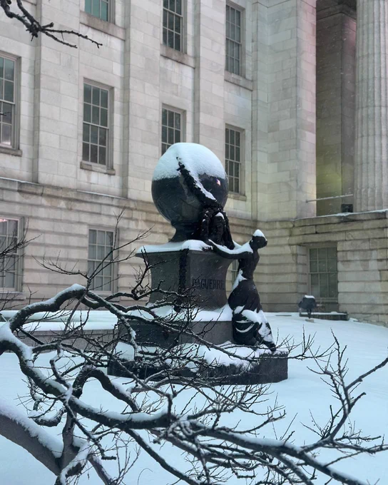 Snow-covered memorial to Daguerre with a large globe and figure of a woman looking up, set against a backdrop of a stone building and branches dusted with snow.