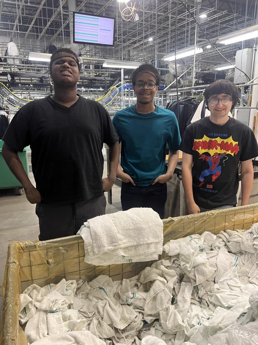 Three young men stand together at a workstation inside a UniFirst facility, smiling toward the camera