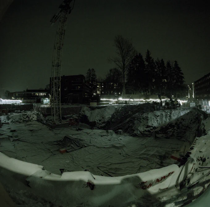 A snow covered building site at night. Big hole in the ground with lights around it.