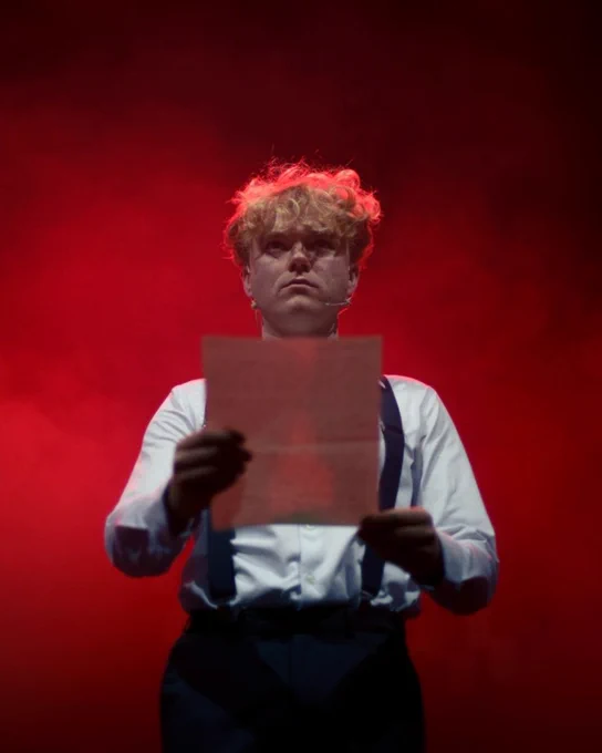 Person holding a sheet of paper on a stage with dramatic red lighting. Photography: Production team