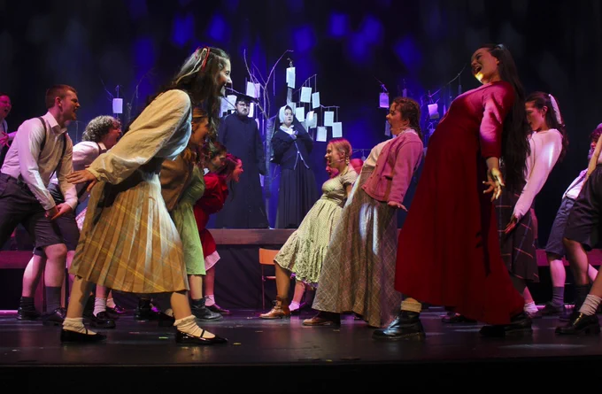 A vibrant stage performance featuring multiple performers in period costumes, dancing and interacting under theatrical lighting with a backdrop of abstract shapes. Photography: Connie Angela McGowan 