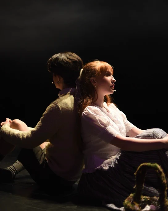 Two actors on stage during a dramatic theater performance, sitting back-to-back with expressive gestures and period costumes. Photography: Production team