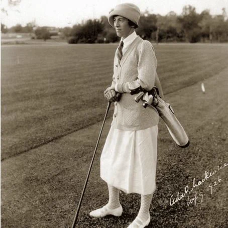 A black-and-white photograph of Ada Mackenzie standing on a golf course. She wears a cloche-style hat, a light cardigan over a blouse, a knee-length skirt, and lace-up shoes. She holds a golf club upright with both hands, with a golf bag resting against her shoulder. The grass fairway stretches behind her with trees in the distance. A handwritten note in the lower right corner reads “Ada Mackenzie” with a date from the early 1920s.
