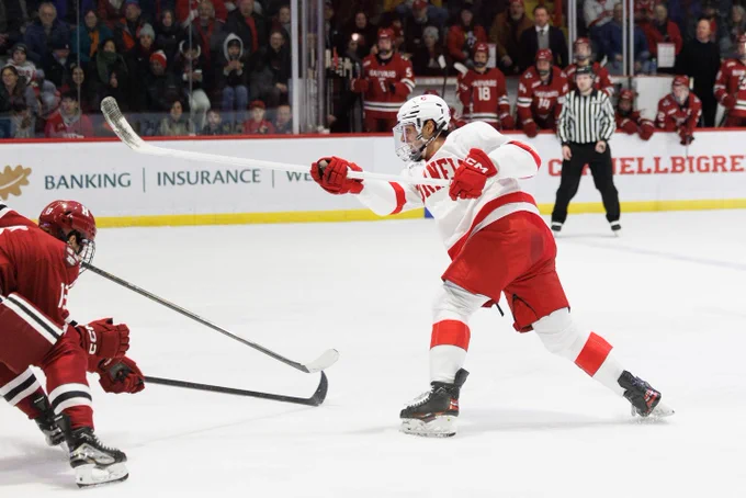 A Cornell University ice hockey player in a white and red uniform maneuvers the puck during a game against a player from Harvard University.