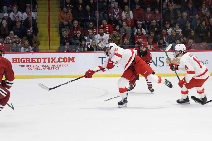 Hockey players from Cornell University and a rival team compete on the ice during a game, with one player taking a shot on goal.