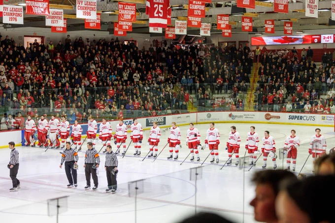 A hockey game between Cornell University and Harvard University, with players lined up on the ice facing each other while referees stand at center ice. The crowded stadium is filled with enthusiastic fans wearing red.
