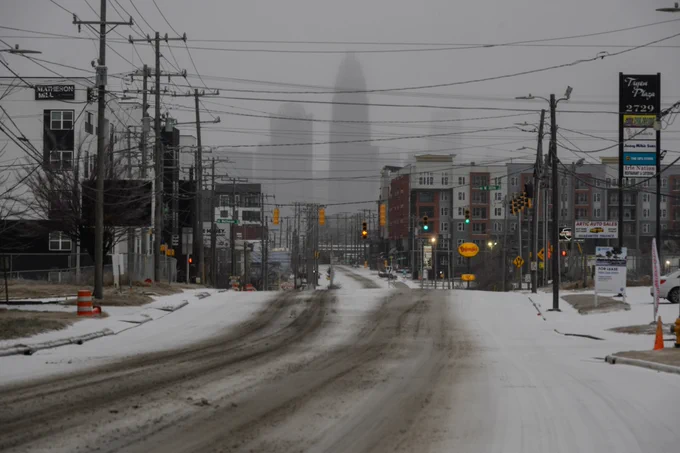 A snowy street scene with traffic lights and road signs, featuring buildings and utility poles lining the road. The Charlotte skyline is partially obscured by fog in the background.