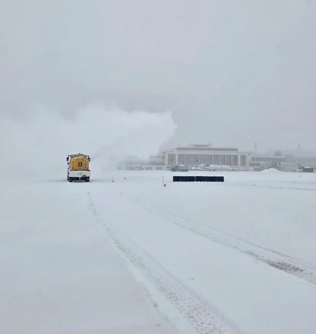 A snowplow clears a runway during a heavy snowfall, with a Reagan National visible in the background.