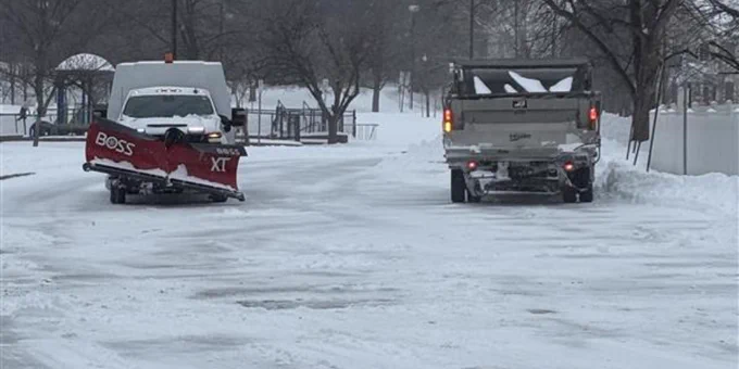 Snow-covered street with a pickup truck and a snowplow clearing the road on a cloudy winter day.