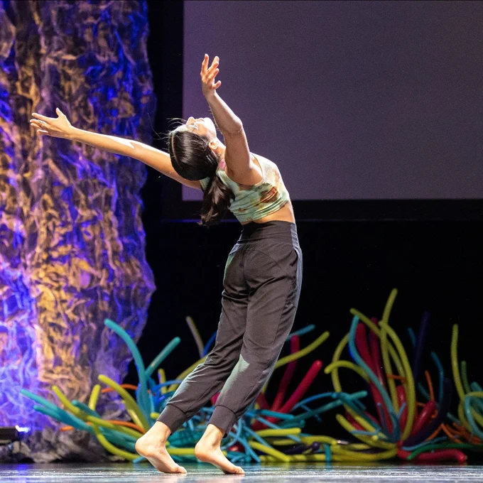 A barefoot dancer arches backward gracefully on stage with arms extended, surrounded by colorful balloon sculptures and textured lighting.