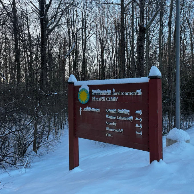A snow-covered wooden sign with the Smithsonian Environmental Research Center logo, at the trailhead of a snowy forest path.