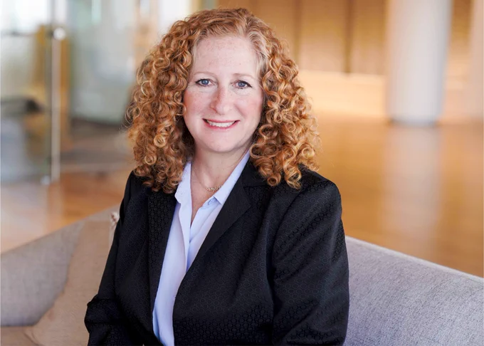 A professional portrait of Jennifer L. Mnookin, a person with curly hair, wearing a black jacket and white shirt, sitting in a modern office environment.