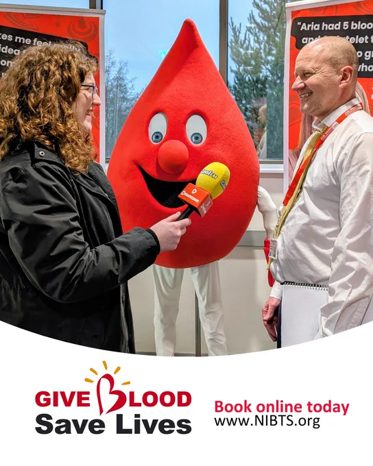 Two people standing smiling in front of a smiling red blood drop mascot costume. A lady on the left holding a microphone interviewing the man on the right. Below the image is text reading 'Give Blood Save Lives' and Book online today www.nibt.org'