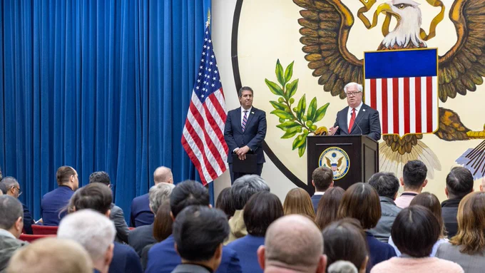 Deputy Secretary Rigas looks at Ambassador Glass who is speaking at audience from a pulpit in front of the state department seal