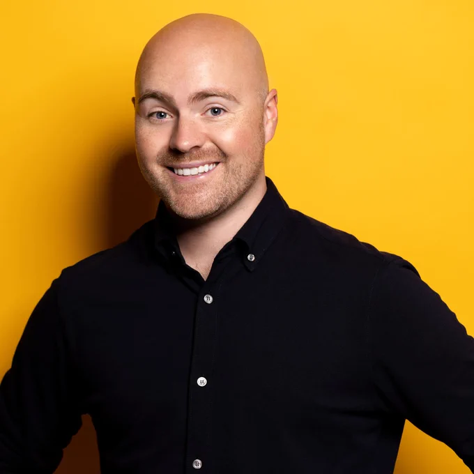 A man stands smiling wearing black short with rolled up sleeves. He stands against a yellow background.