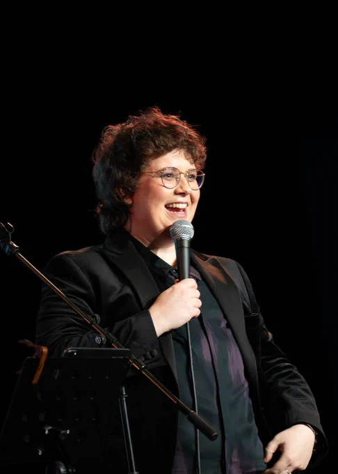 A person stands smiling and talking into a microphone. They wear black and the background is dark. 