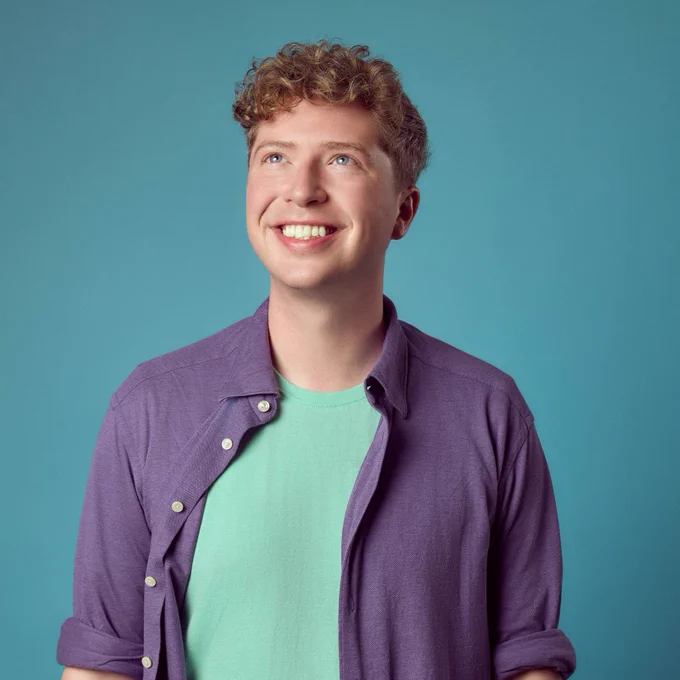 A man looks upwards smiling. He wears a purple shirt with rolled up sleeves, a mint t-shirt and aubergine trousers. The background is blue. 