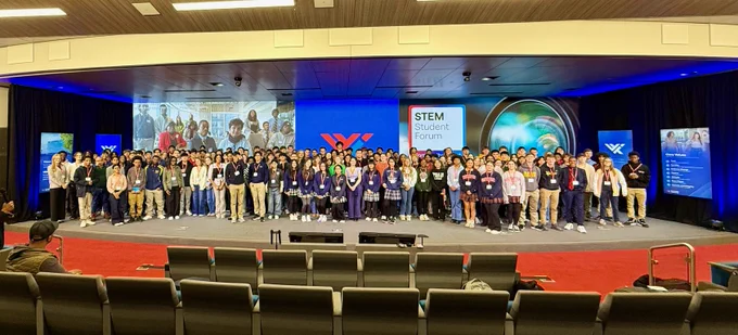 A wide shot of a large group of students on stage at the STEM Student Forum, with screens displaying related content and the event logo in the background. Audience chairs are visible in the foreground.