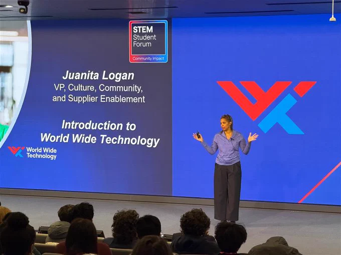 Juanita Logan, VP at World Wide Technology, presents to an audience. A screen behind her displays her name, title, and the company logo. Text reads: STEM Student Forum, Community Impact, and Introduction to World Wide Technology.
