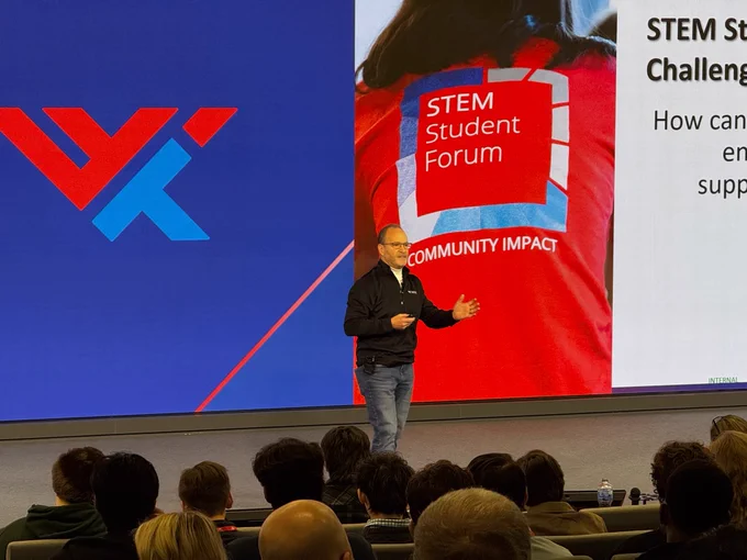 A man is giving a presentation at a STEM student forum. The screen displays the organization's logo in red and blue, along with the text \"STEM Student Forum\". He is wearing a black jacket and jeans.
