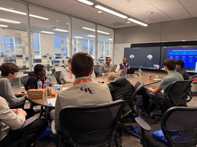 A group of people are gathered around a conference table, some looking towards the front, where there are two screens displaying \"Global Operations Supply Chain Service\". The back of a jacket with \"CBC\" on it is visible.