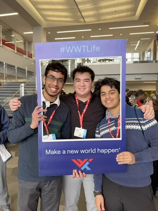 Three smiling young men pose inside a blue frame that reads \"#WWTLife\" and \"Make a new world happen\". 