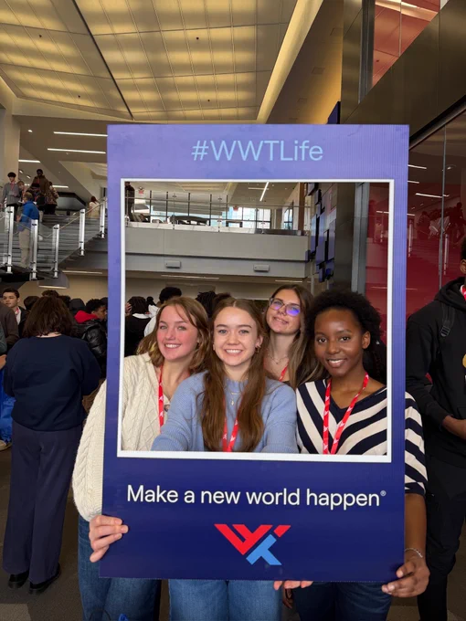 Four smiling young women pose inside a large frame with \"#WWTLife\" at the top and \"Make a new world happen\" with a logo at the bottom. Stairs and a crowd are in the background.