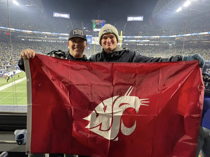 Two individuals stand together inside a football stadium, holding a large red Washington State University flag featuring the white Cougar logo. The brightly lit field and packed stands are visible behind them during a nighttime game.