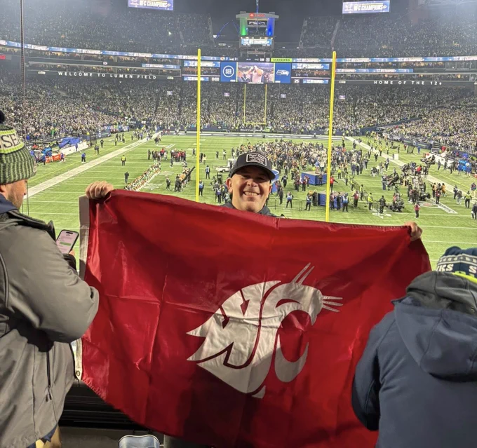 A person stands in a stadium overlooking a crowded football field at night, holding up a large red Washington State University flag with the white Cougar logo. Fans fill the stands, and activity is happening on the field below under bright stadium lights.