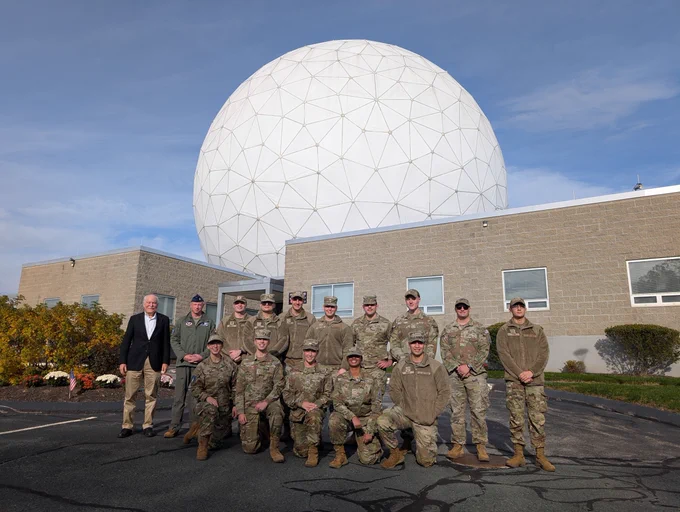 Group of military personnel and two civilians posing in front of a large geodesic radar dome on a clear day.