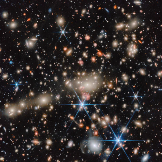 A Webb image of many glowing galaxies in deep space, in various shapes and colours, on a black background. There are some large, blue spiral galaxies, some large and pale white elliptical galaxies, and many orange and red, medium-sized galaxies. Even smaller galaxies, down to tiny faint spots, appear in all these colours.