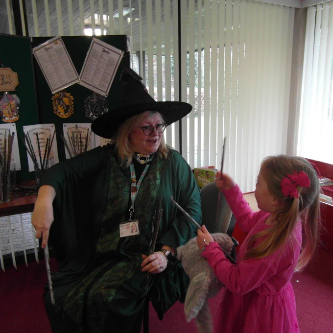 A library staff member wears a witch hat and looks at a young girl in a pink top who is choosing a magic wand