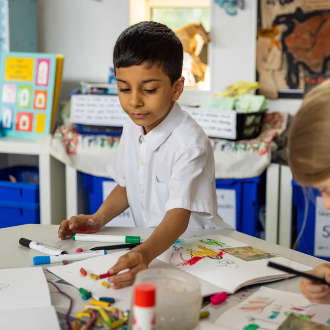 A child in a white shirt draws colorful pictures with markers and crayons at a classroom table filled with art supplies.