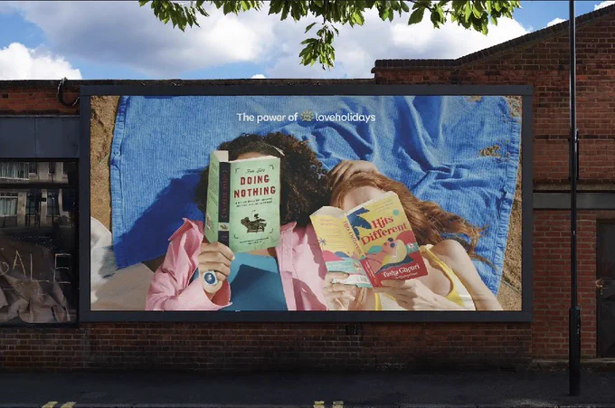 A billboard showing a photo of two people lying down, books in front of their face. The books are Doing Nothing by Tom Lutz and Hits Different by Tasha Ghouri & Lizzie Huxley-Jones, spelling out “doing nothing hits different”. The billboard is a campaign for LoveHolidays.