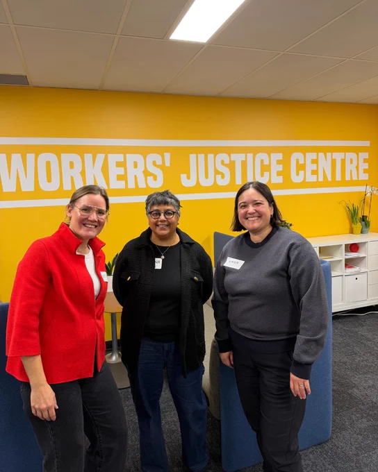 Photo of three people posing and smiling in front of a wall that reads “Worker’ Justice Centre”.  