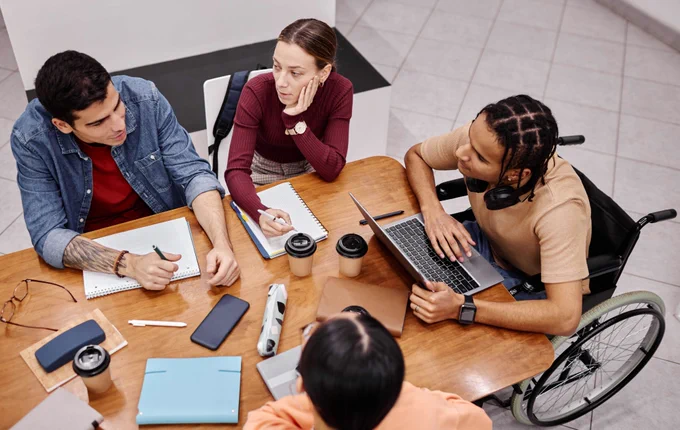 Group of students of various sexes and races, including a person in a wheelchair, sitting around a conference table. 