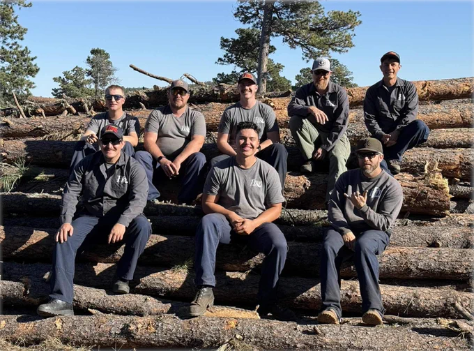 Wildland firefighters sitting on stacked logs under a clear blue sky in a forested area.