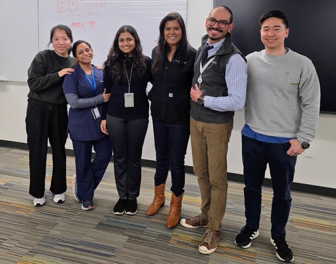 Six professionals smiling in a modern office setting, with a whiteboard in the background showing some written content.
