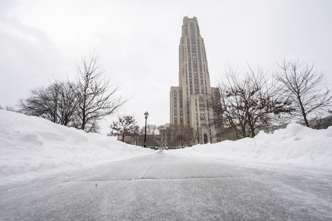 Low angle view of a snow-covered path leading to the Cathedral of Learning at the University of Pittsburgh, flanked by bare trees.