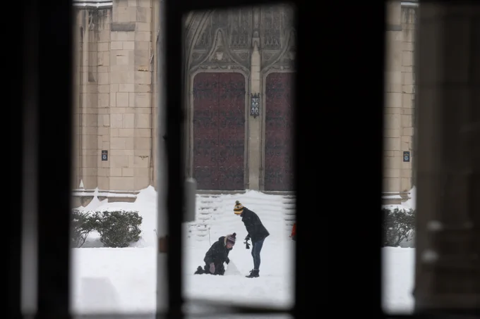 View through an archway showing two people playing in the snow in front of a historic building with large ornate doors.
