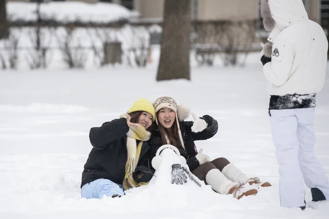 Two individuals sitting in snow, smiling and posing for a photo with a small snowman, while another person stands nearby.
