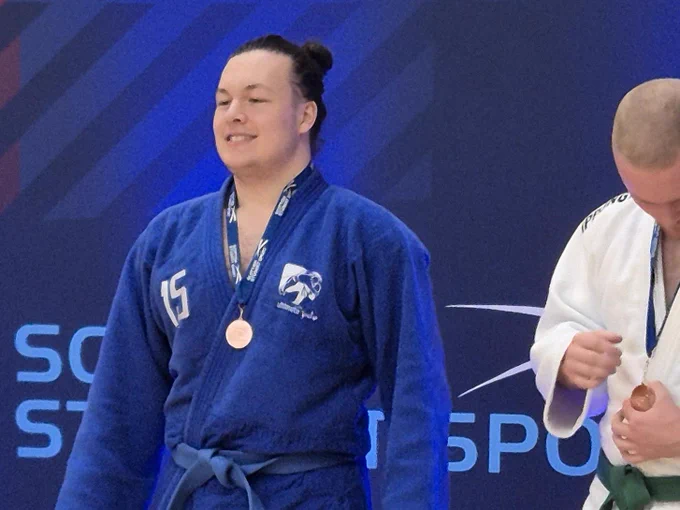 Barry Shum, wearing a blue judo gi, stands wearing a bronze medal, smiling during a Scottish Student Sport judo competition. Another competitor is partially visible beside him against a branded event backdrop.