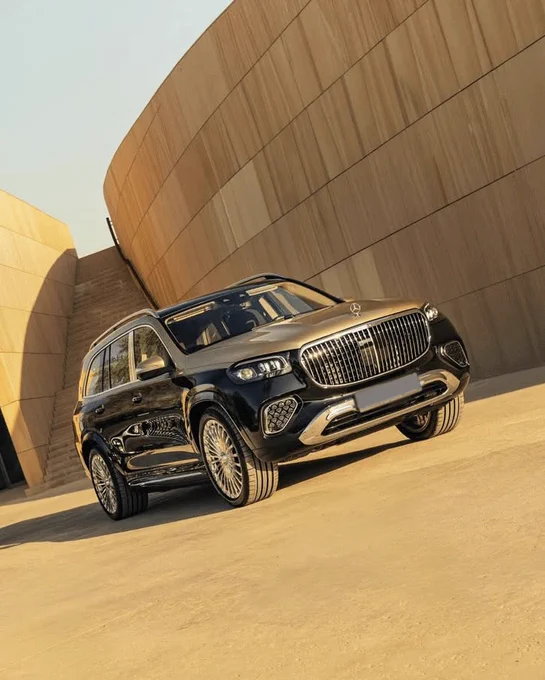 A Mercedes-Maybach GLS parked on a smooth, sandy surface. The vehicle has a sleek design with a prominent grille and shiny wheels. The background features a modern architectural structure with wooden textures and curved lines.
