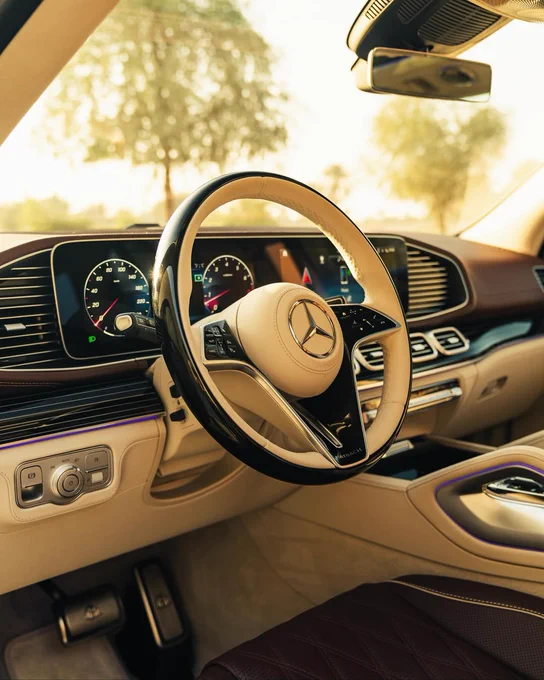 Interior of a luxury car, featuring a Mercedes-Benz steering wheel, digital dashboard with dials and displays, leather seating, and detailed controls. The sunlit exterior is visible through the windshield, adding a warm glow to the scene.