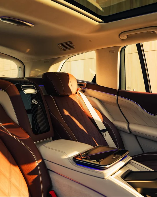Interior of a luxury car, featuring plush brown and white leather upholstery. The back seats are equipped with a central console, containing a touchscreen interface. Soft lighting and windows illuminate the spacious cabin.