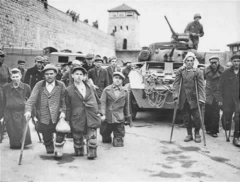 A black and white photo of a crowd of wounded war veterans and disabled people gathered around an army tank. Tall walls enclose the space and there is a guard tower visible in the background.
