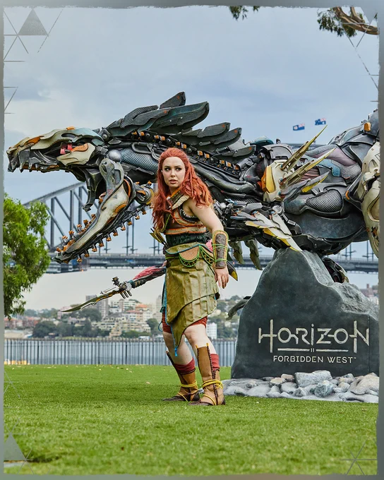 Photo of an Aloy cosplayer wearing the Utaru Harvester armor posing in front of a large statue of a Clawstrider.